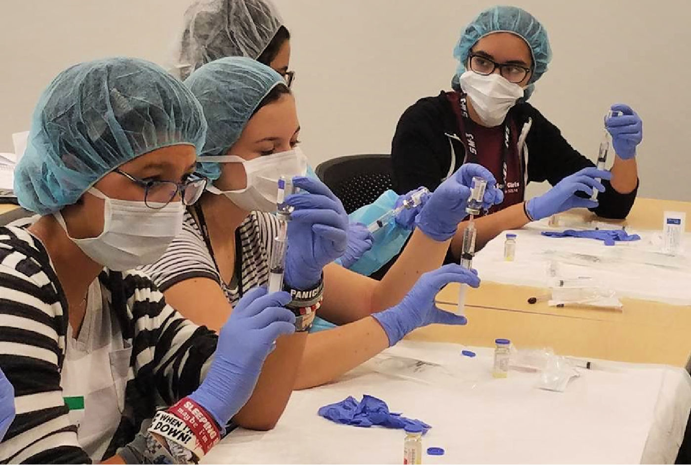 A group of four students in medical attire focus on handling syringes and vials during a training session.