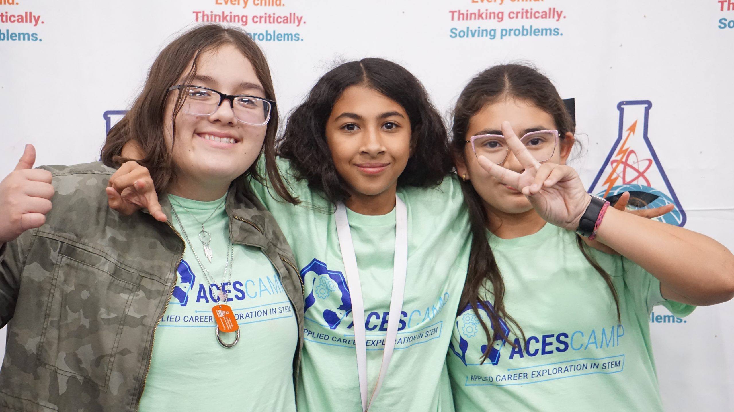 Three girls standing in front of a SARSEF photo banner smiling at the camera. Each girl is wearing a light green ACES camp shirt.